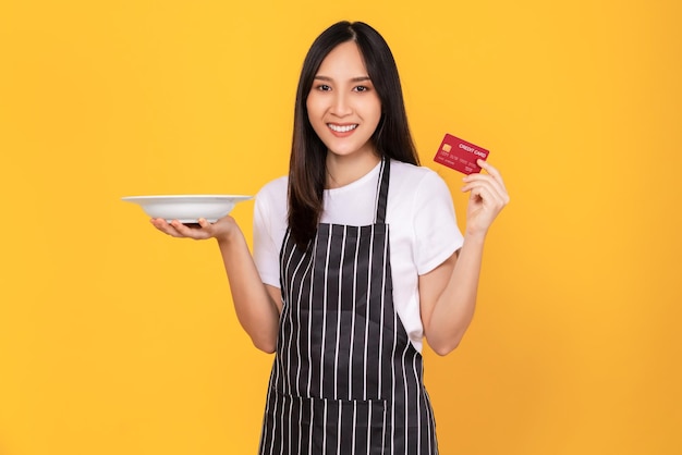 Foto le belle donne asiatiche indossano il grembiule con in mano piatti bianchi e mostrano la carta di credito su sfondo giallo.