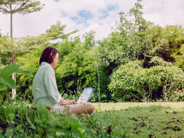 緑の芝生に座っているラップトップコンピューターで白いシャツとショートパンツを着ている美しいアジアの女性。