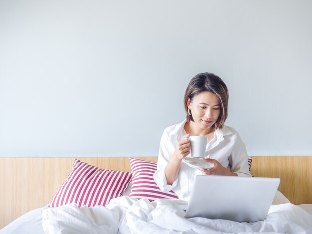 Beautiful Asian women with short hair wearing white shirt working with laptop computer on the bed in the house.