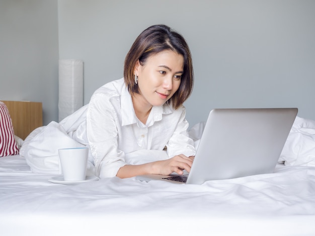 Belle donne asiatiche con la camicia bianca d'uso dei capelli corti che lavora con il computer portatile sul letto nella casa.