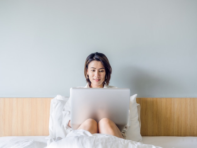 집에서 침대에 랩톱 컴퓨터를 사용하는 흰색 셔츠를 입고 짧은 머리를 가진 아름 다운 아시아 여자.
