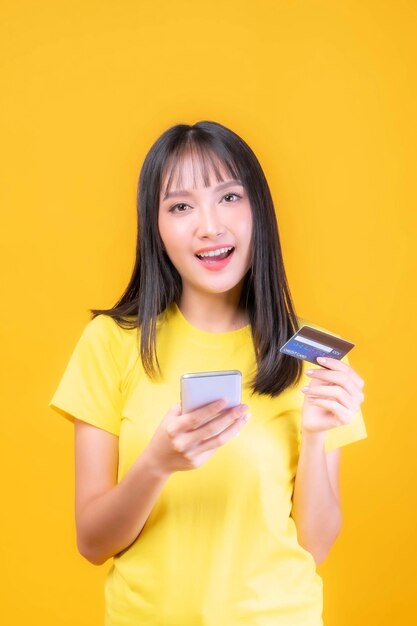 Beautiful Asian young woman cute girl with bangs hair style in yellow shirt holding credit card and smartphone ,mobile phone uses a online banking for pay online shopping isolated on yellow background