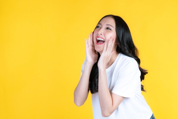 Beautiful Asian young woman screams announce the good news or promotion , holding hands near her face with open mouth Herald news promotion isolated on yellow background