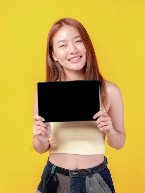 Foto bella giovane asiatica che mostra smartphone tablet computer con schermo vuoto schermo nero spazio di copia vuoto per banner pubblicitario isolato su sullo sfondo giallo mock up image