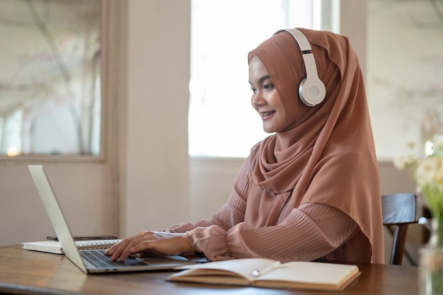 ヒジャブとヘッドフォンを着た美しいアジア系イスラム教徒の女性が ⁇ カフェでラップトップで作業しています ⁇ 