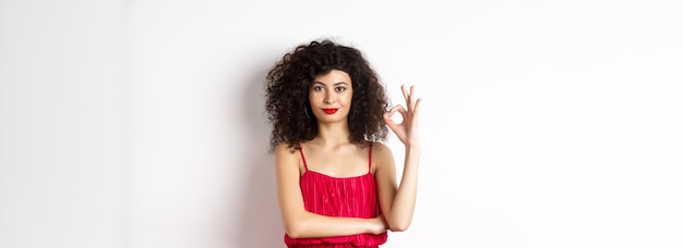 Photo beautiful assertive woman with curly hairstyle and makeup wearing red dress showing okay sign and sm