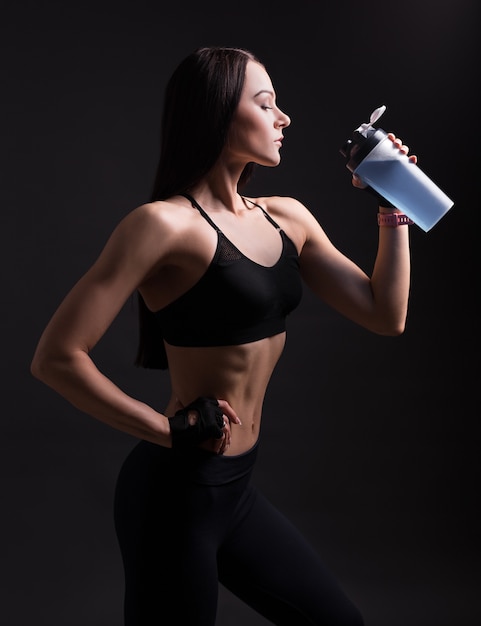 Beautiful athletic woman in sportswear drinking protein cocktail over black background