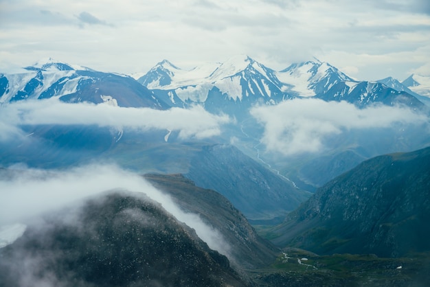 Фото Красивый атмосферный альпийский пейзаж с большими заснеженными горами с ледником. низкие облака среди скал в зеленой долине с речкой. замечательные горные пейзажи. полет над горами над облаками.