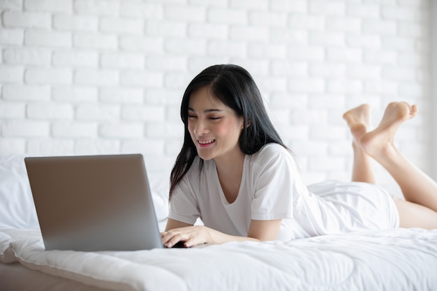 Foto bella giovane donna asiatica attraente che si trova sul letto e che utilizza il computer portatile del computer per l'utilizzo dei media sociali