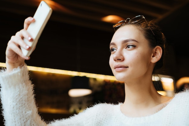 Beautiful attractive girl resting in a cozy cafe and making selfie on her smartphone