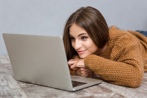 Beautiful attractive happy relaxed young woman in brown sweater lying on wooden floor using laptop