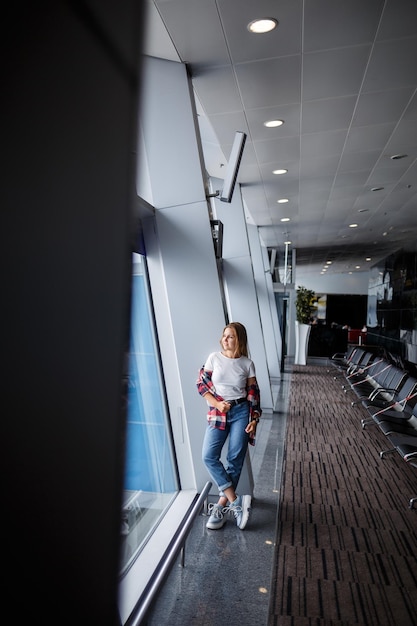 A beautiful attractive ukrainian woman with blond hair is standing by a large window wearing momom jeans and a tshirt with a plaid shirt Girl in casual comfortable clothes