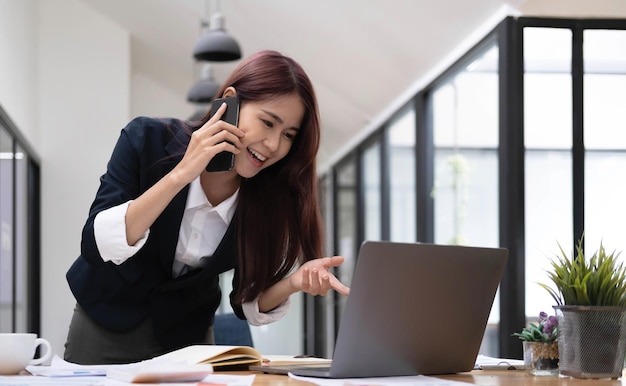 Beautiful attractive young asian businesswoman talking on the phone with her colleague and checking the informations on laptop screen