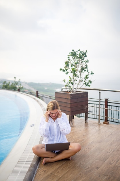 Beautiful attractive young woman sits near a large pool and works at a laptop. Remote work while on vacation.