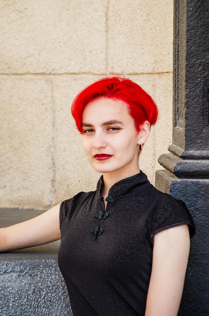 Foto bella giovane donna attraente con capelli rosa in abito nero si siede sullo sfondo del muro di pietra