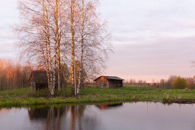 Beautiful autumn or spring landscape, old log houses on the shore of a pond or lake in the shade of tall birches.