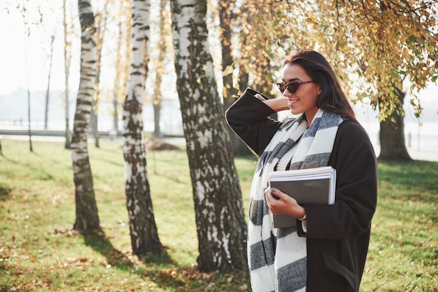 Beautiful autumn. Young smiling brunette in sunglasses stands in the park near the trees and holds notepad