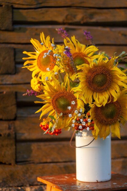 田舎の木製のテーブルに白い花瓶に明るい黄色のヒマワリの花の美しい秋の花束。庭の花のある秋の静物。