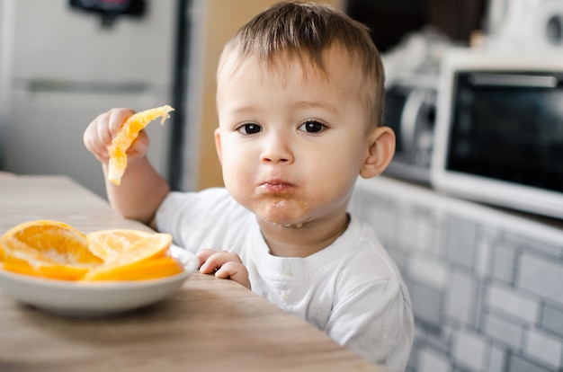 Bellissimo bambino in cucina che mangia avidamente un'arancia, tagliata a spicchi