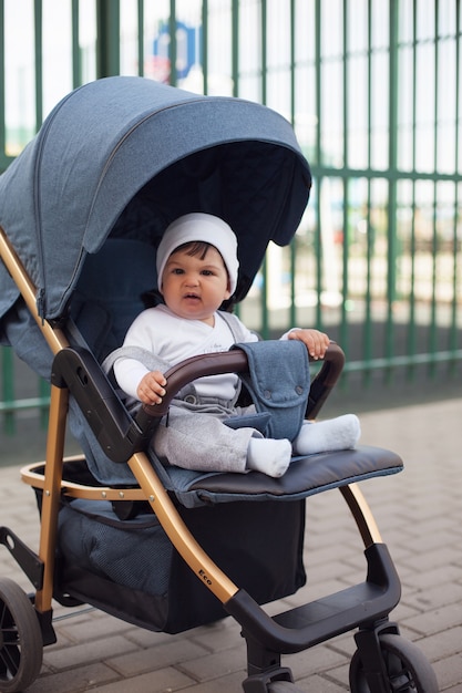 A beautiful baby boy in a white cap sits in a baby carriage, smiles and wrinkles his nose