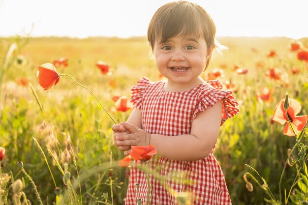 Beautiful baby girl in red dress on field of poppies at summer sunset.