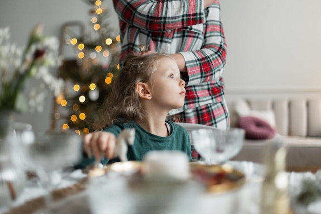 Beautiful baby girl sits at the table and mom braids her hair on Christmas Eve Family winter holiday Waiting for a miracle