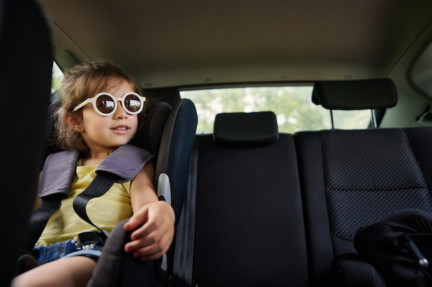 サングラスをかけた美しい女の赤ちゃんが車のチャイルドシートに座って窓の外を見ています。車内で子供と安全に旅行