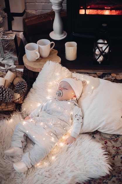 Beautiful baby sleeps on the coverlet with a lot of lights and gifts around him near the electric fireplace