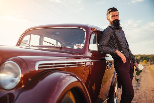 Beautiful bearded brunette man in a shirt, trousers with suspenders holds a white rose in his hand near a brown retro car on a country road.