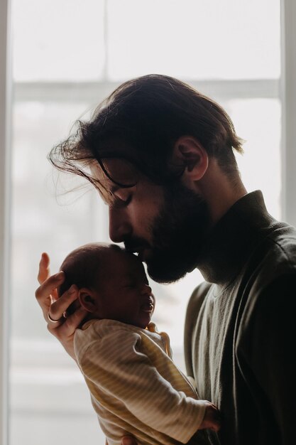 Beautiful bearded father kisses newborn daughter in a dark room near the window