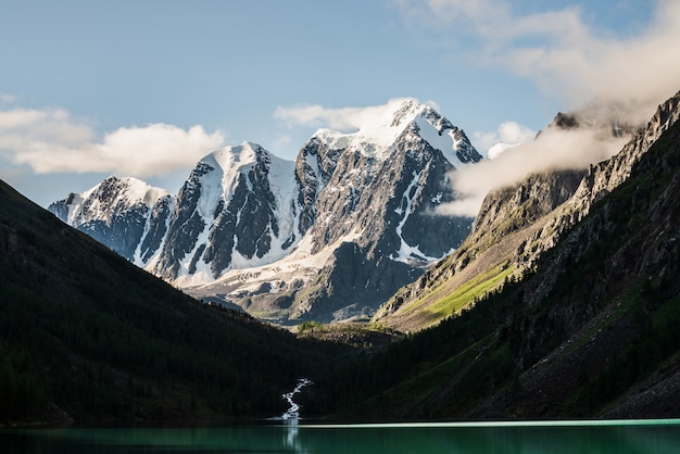 Beautiful big glacier, rocky snowy mountains, coniferous forest on hills, mountain lake and highland creek under blue sky with clouds.