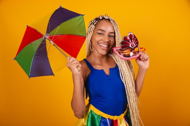 Beautiful black Brazilian woman dressed in carnival clothes blue and yellow background holding umbrella and mask