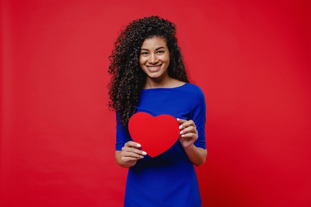 Beautiful black woman in blue dress with heart shaped valentine card on red wall