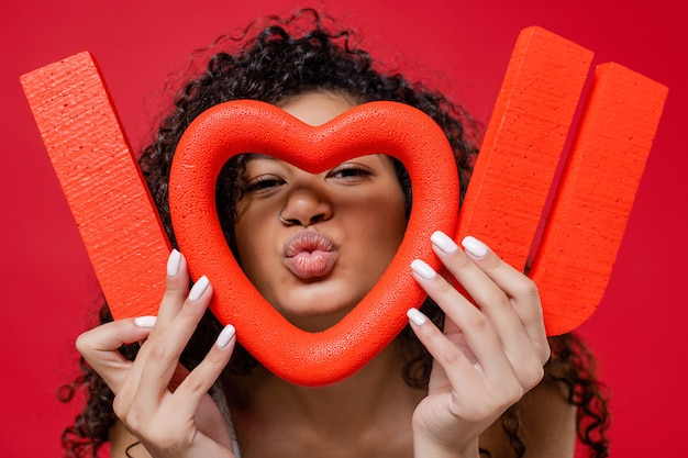 Beautiful black woman holding i love you letters looking through heart shape isolated on red