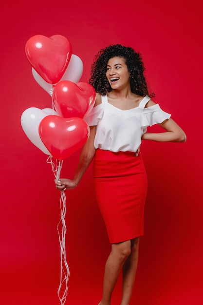 Beautiful black woman smiling with heart shaped colorful balloons on red wall