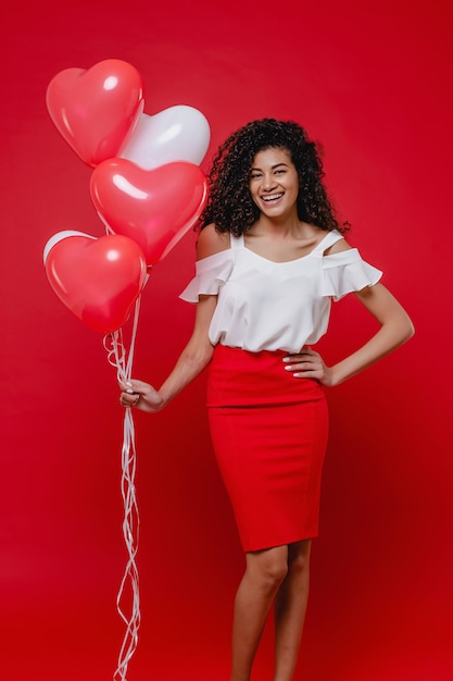 Beautiful black woman smiling with heart shaped colorful balloons on red wall