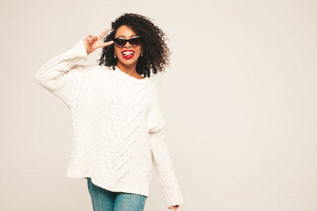 Beautiful black woman with afro curls hairstyle and red lips.Smiling model in  trendy jeans clothes and winter sweater. 