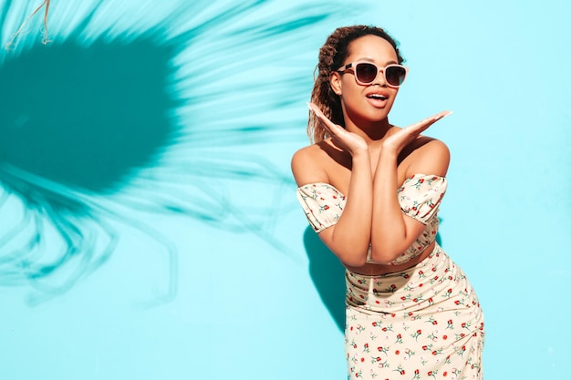 Foto bella donna di colore con l'acconciatura di riccioli afro modello sorridente vestito con abiti estivi hipster donna sexy spensierata in posa vicino al muro blu in studio abbronzato e allegro in occhiali da sole