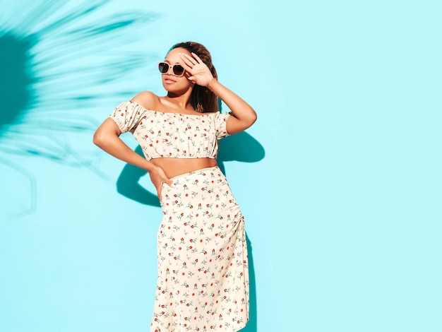 Photo beautiful black woman with afro curls hairstyle smiling model dressed in summer hipster clothes sexy carefree female posing near blue wall in studio tanned and cheerful in sunglasses