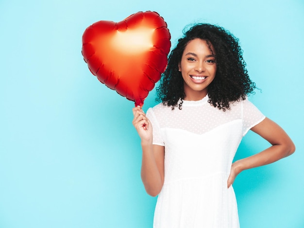 Beautiful black woman with afro curls hairstyle Smiling model dressed in white summer dress Sexy carefree female posing near blue wall in studio Tanned and cheerful Holding heart air balloon