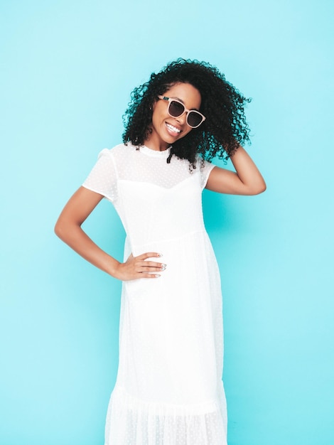 Beautiful black woman with afro curls hairstyle Smiling model dressed in white summer dress Sexy carefree female posing near blue wall in studio Tanned and cheerful in sunglasses Isolated