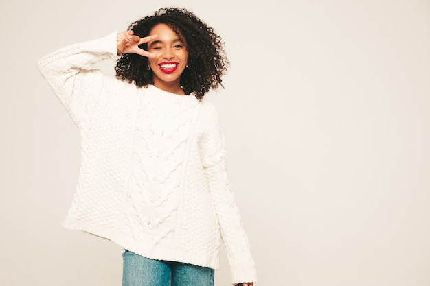 アフロカールの髪型を持つ美しい黒人女性。白い冬のセーターとジーンズの服の笑顔モデル。