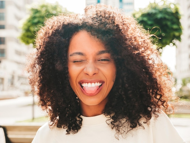 Beautiful black woman with afro curls hairstylesmiling hipster model in white tshirt sexy carefree female posing on the street background cheerful and happyshows tongue