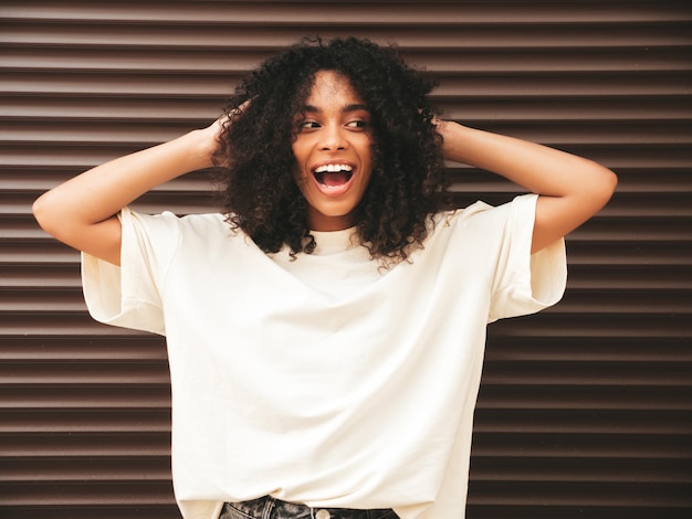 アフロカールの髪型を持つ美しい黒人女性白いTシャツの笑顔のヒップスターモデル茶色の壁の近くの通りでポーズをとるセクシーな屈託のない女性陽気で幸せ