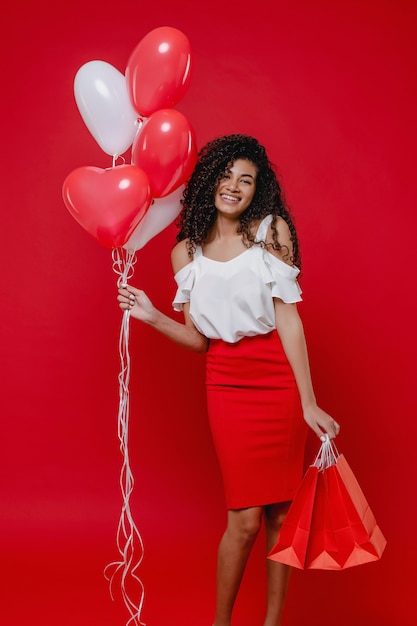 Beautiful black woman with colorful heart shaped balloons and shopping bags on red wall