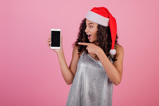 ピンクの上に分離されたクリスマス帽子をかぶっている電話のcopyspace画面と美しい黒人女性