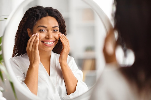 Beautiful black young woman looking in mirror and applying under eye cream