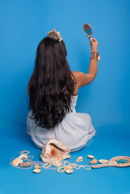 Photo beautiful blackhaired woman wearing white clothing sitting against blue background next to several sea shells with her back holding and looking into a mirror