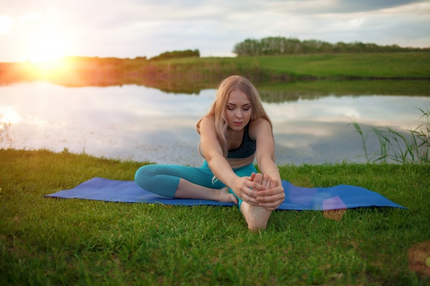 Красивая белокурая девушка практикует йогу на озере на закате. крупный план поддерживает здоровый образ жизни