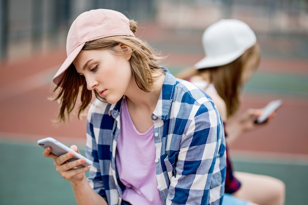 市松模様のシャツとキャップを身に着けている美しいブロンドの女の子が電話でスポーツフィールドに座っています